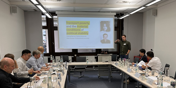 Auf dem Bild ist ein Konferenzraum, in dem die Workshopteilnehmer*innen an Tischen sitzen und einem Vortrag folgen. In der Mitte ist eine Leinwand, auf dem Folien des Vortrags zu sehen sind.