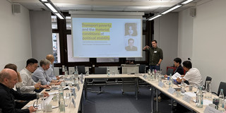 Auf dem Bild ist ein Konferenzraum, in dem die Workshopteilnehmer*innen an Tischen sitzen und einem Vortrag folgen. In der Mitte ist eine Leinwand, auf dem Folien des Vortrags zu sehen sind.