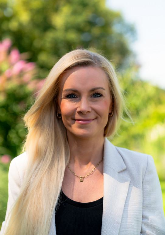 Auf dem Bild ist Meike Neuhaus. Sie hat blonde Haare, trägt ein helles Jacket und ein schwarzes Shirt. Sie steht draußen vor einem verwaschenen grünlichen Hintergrund.