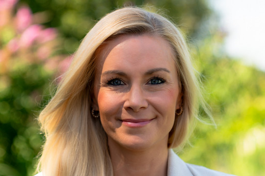 Auf dem Bild ist Meike Neuhaus. Sie trägt ein helles Jackett und ein schwarzes Shirt. Sie steht draußen vor einem verwaschenen grünlichen Hintergrund.