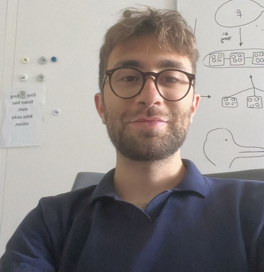 Auf dem Bild ist Riccardo Fracchioni. Er trägt ein dunkles T--Shirt und sitzt vor einer weissen Wand.