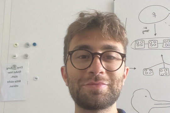 Das Bild zeigt Riccardo Fracchioni. Er trägt ein dunkles T-Shirt und sitzt vor einer wissen Wand.