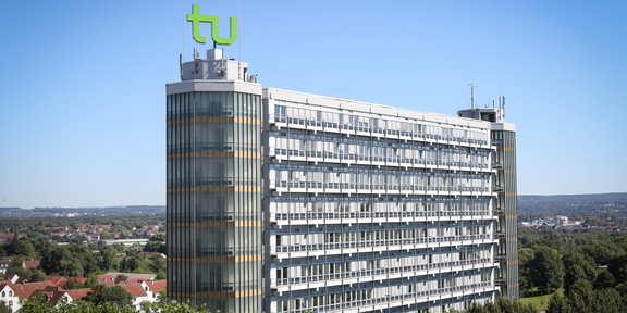Mathematikgebäude mit Mathetower: Ein hohes Gebäude mit vielen Fenstern und je einem Turm an beiden Seiten. Auf dem linken Turm ein grünes TU-Logo. Im Hintergrund Häuser und Bäume.
